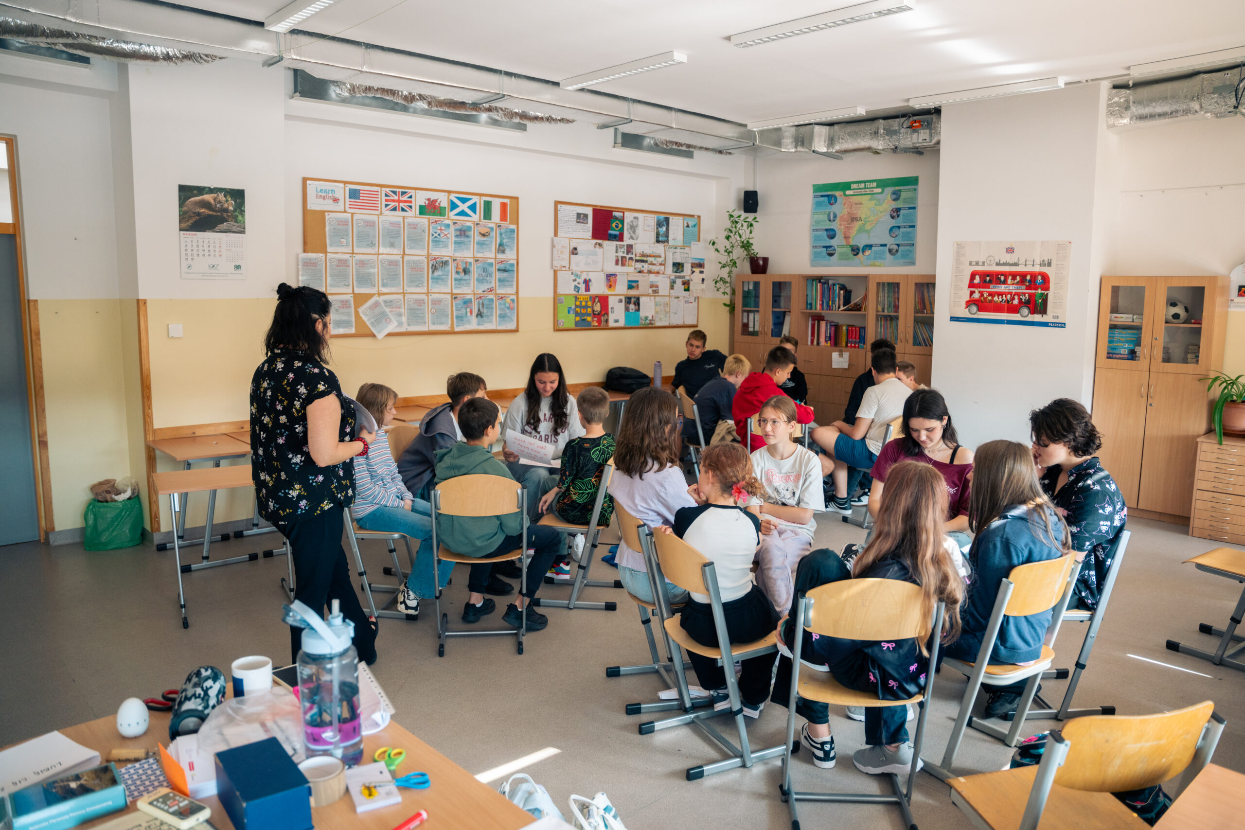 Obrazek przedstawia prezes Fundacji A.R.T. - Agnieszkę Tobota, która przygląda się pracy uczniów, w grupach - okręgi ustawione z krzesełek ze stolikami pośrodku.