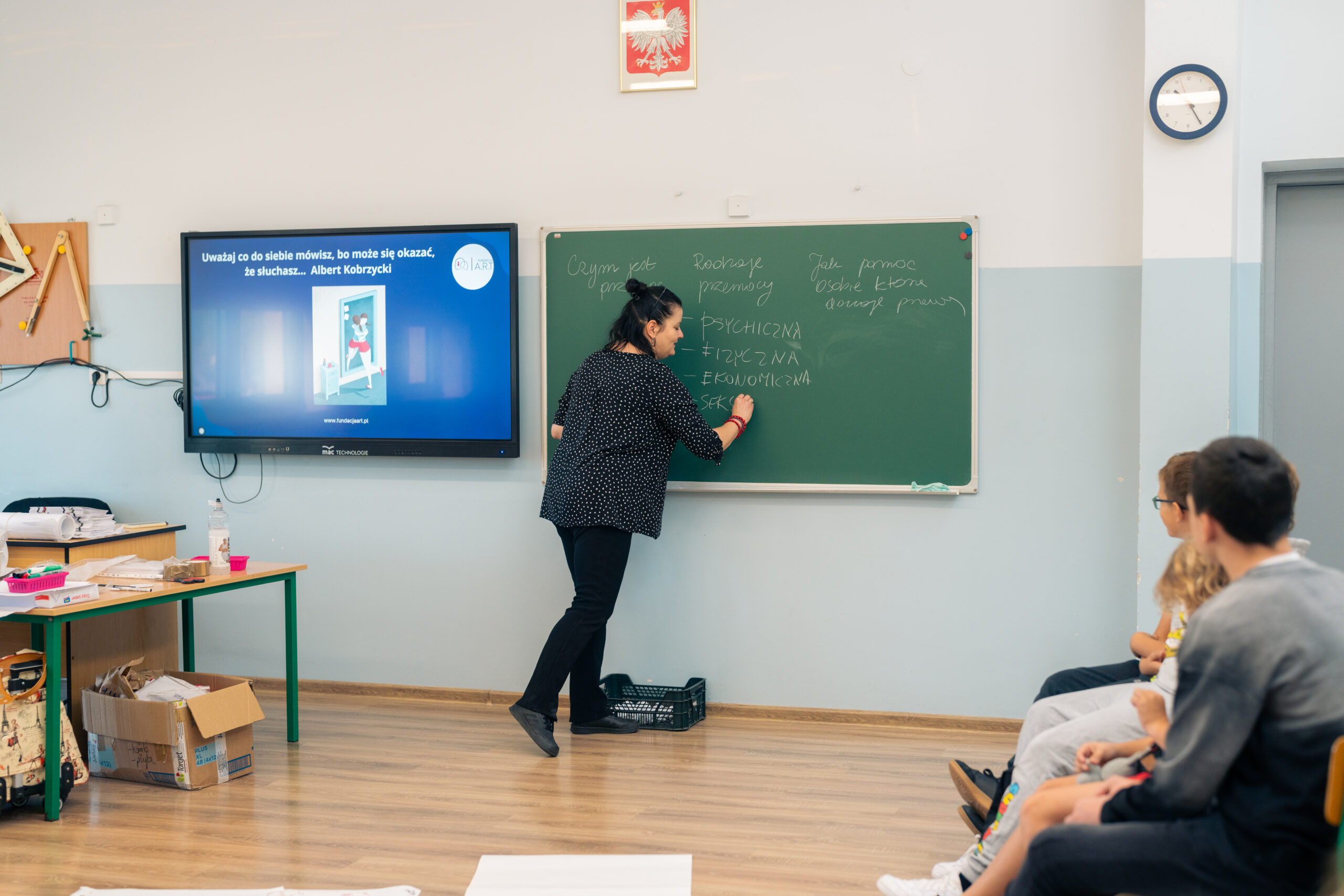 Obrazek przedstawia prezeskę Fundacji A.R.T. - Agnieszkę Tobota w klasie lekcyjnej. Zapisuje słowa na zielonej tablicy kredą.