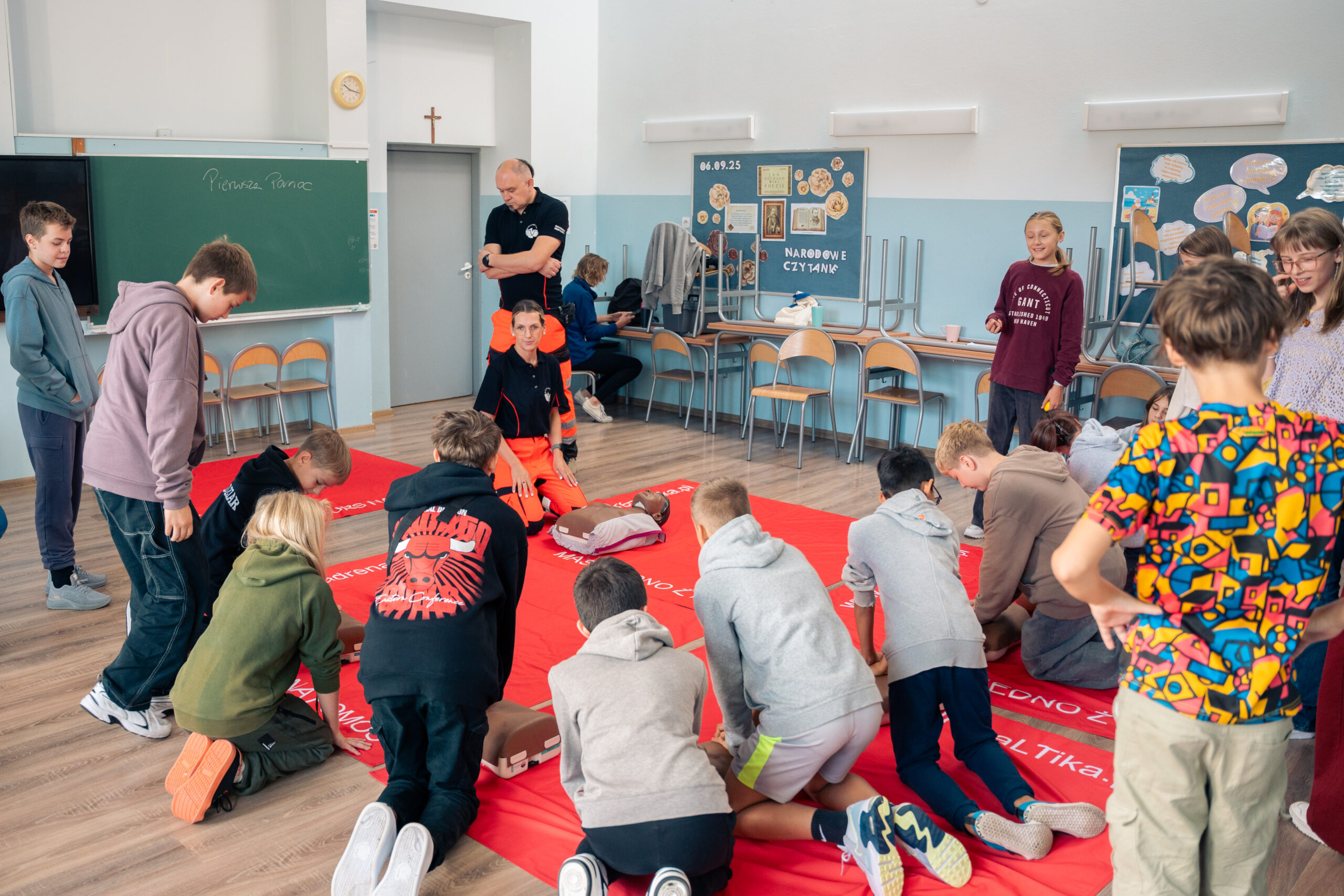 Obrazek przedstawia ratowników medycznych demonstrujących jak wykonywać RKO na manekinach oraz uczniów, którzy również wykonują RKO na manekinach, idąc za przykładem ratowników.